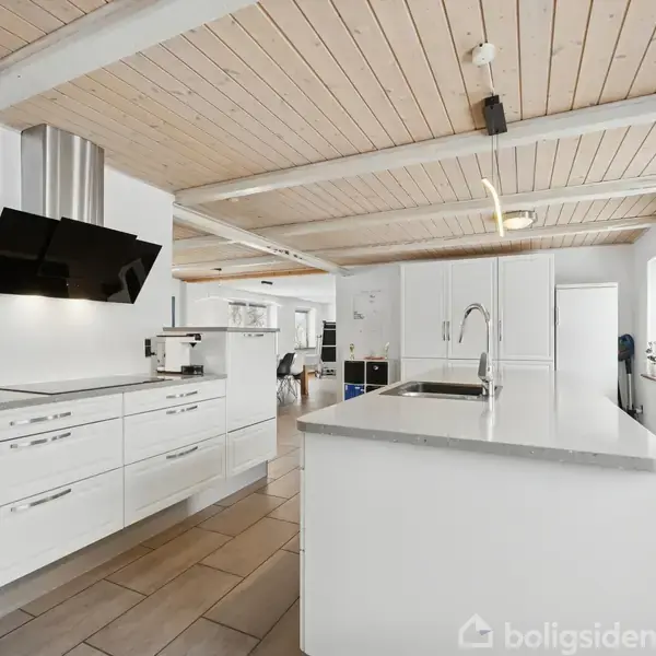 Lyst køkken med hvide skabe og en central køkkenø med integreret vask, beliggende i en moderne bolig. Åben planløsning fører til spisestue med spisebord i baggrunden.