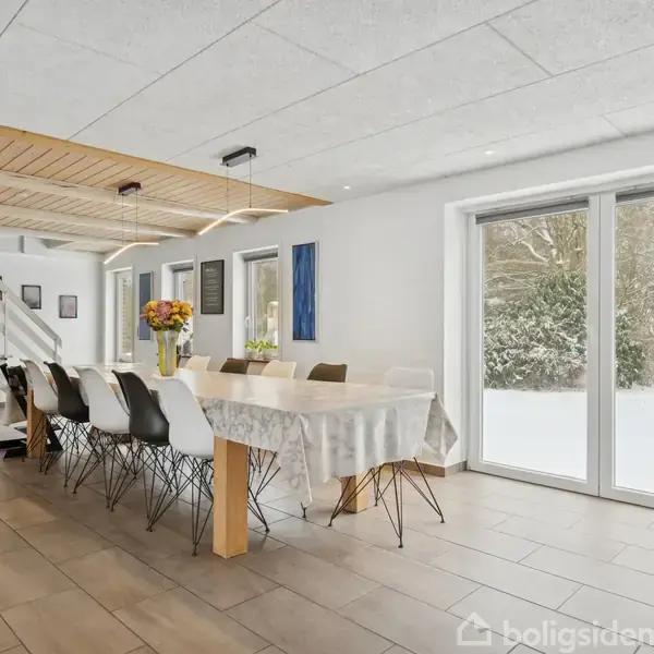 Et langt spisebord dækket med en dug står i et lyst rum med store vinduer, der viser en snedækket have. Vasen med blomster pynter bordet, mens trapper fører opad.