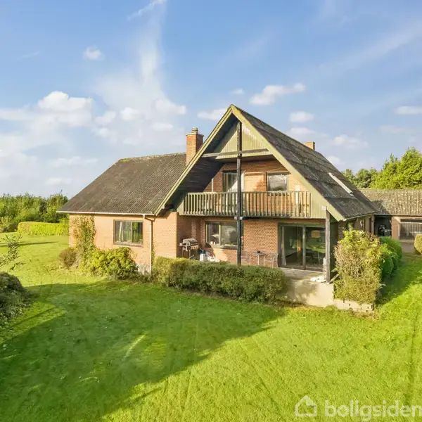 Et stort, trekantet hus med en balkon er omgivet af en velplejet græsplæne og omgivet af buske og træer under en blå himmel.