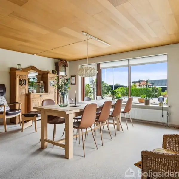 Langbord med otte stole står i en lys spisestue. Rummet har store vinduer med udsigt til en have samt dekorerede vægge med planter, trofæer og en skænk.