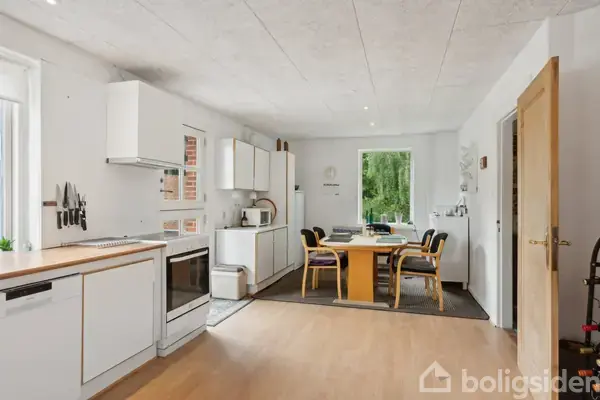 Et lyst køkken med hvide skabe og ovn til venstre. Et spisebord med seks stole står i midten, omgivet af vinduer med udsigt til en grøn have.