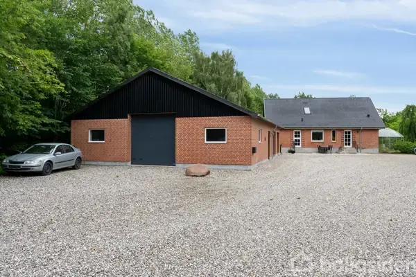 En rød murstensbygning med sort tag står stille på en grusbelagt plads, omgivet af frodig grøn vegetation og en parkeret bil på venstre side.