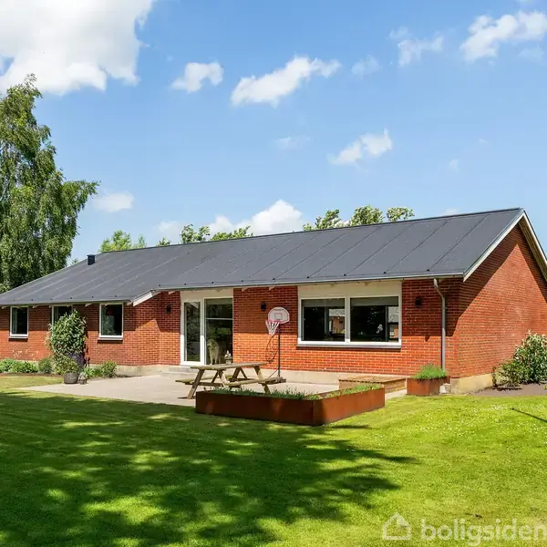 En rød murstensvilla med sort tag ligger i grøn have. Et picnicbord står foran huset. Et drivhus er synligt til venstre, omgivet af træer og plæner.