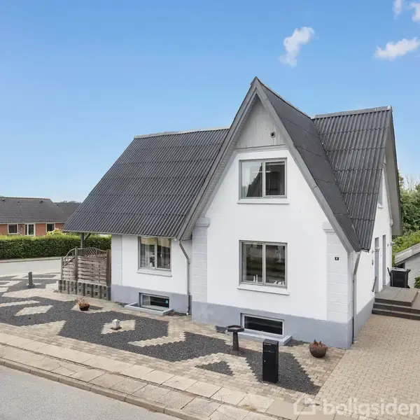 Et hvidt hus med sort tag står på en stille gade. Huset har flere vinduer og et indgangsparti med fliser. Der er en lille have og andre huse i nabolaget.