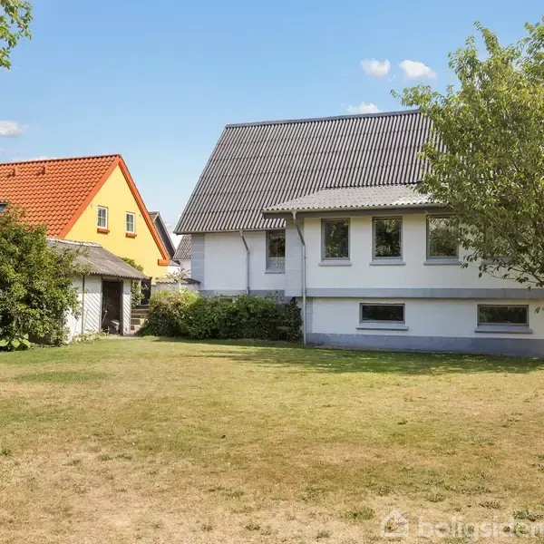 Et hvidt hus med skråt tag står i en have med græs. Til venstre ses et hus med orange tag. Træer giver skygge og omkranser grunden.