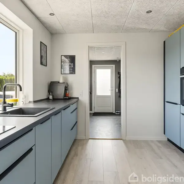 Køkken med lyseblå skabe og sorte bordplader; der er en vask ved vinduet. I baggrunden leder en dør til en lys gang. Vægge er hvide med billeder.