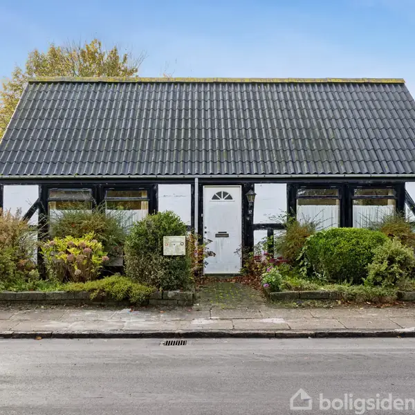 Et hvidt bindingsværkshus med sort tag står i en gade omgivet af buske og træer. Huset har tre store vinduer og en hvid dør. Tilsyneladende en rolig boligområde.