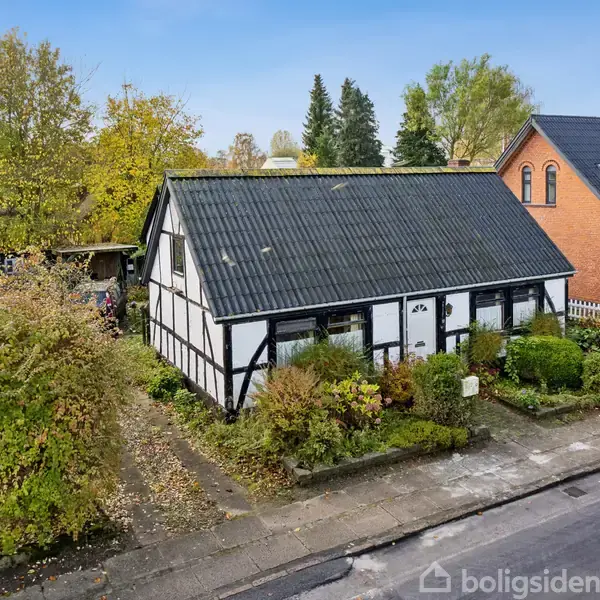 Bindingsværkshus med sort tag står stille i en frodig have, omgivet af buske og træer, ved siden af et rødt murstenshus under en blå himmel.