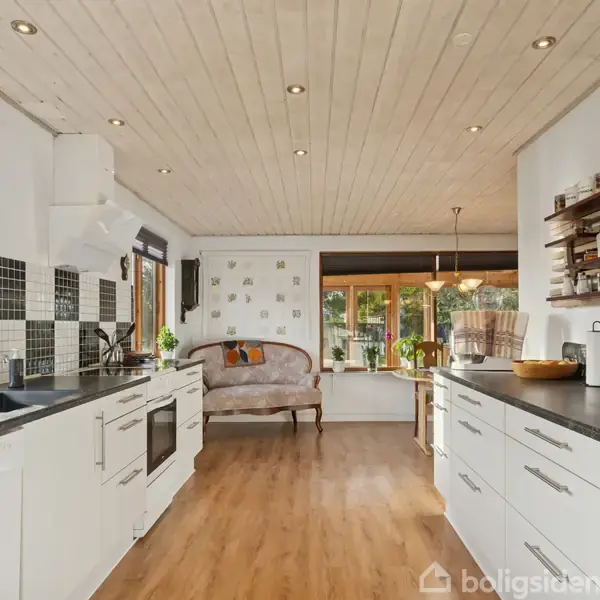 Et lyst køkken med hvide skabe og sorte bordplader strækker sig langs væggene. Midt i rummet er der en sofa, mens store vinduer giver udsigt til en grøn have.