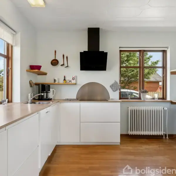 Et lyst køkken med hvide skabe og træbordplader. Køkkenredskaber hænger på væggen. Mørk emhætte over komfuret. Store vinduer giver udsigt til en have.