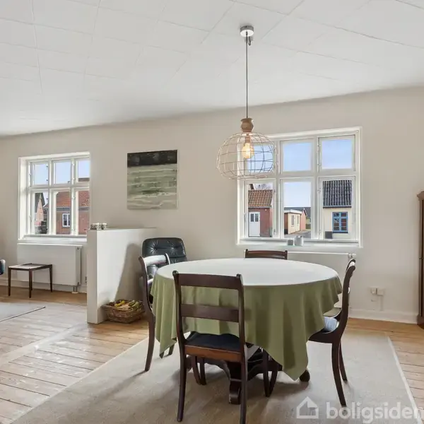 Et rundt spisebord dækket med en grøn dug står i en lys stue med træmøbler. To vinduer giver udsigt til hustage udenfor. Der hænger en enkelt lampe over bordet.