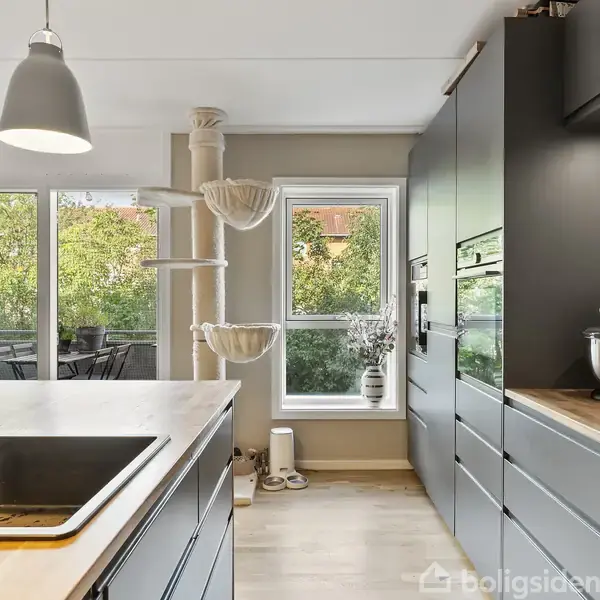 Et moderne køkken med sorte skabe og træbordplade. En stor vask med armatur er i forgrunden. Mod baggrunden ses et kattetræ, en terrasse og grønne omgivelser udenfor vinduet.