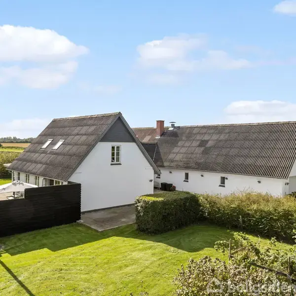 Et hvidt hus med skråt tag står stille på en grøn græsplæne omgivet af hække og træer under en blå himmel med få skyer.
