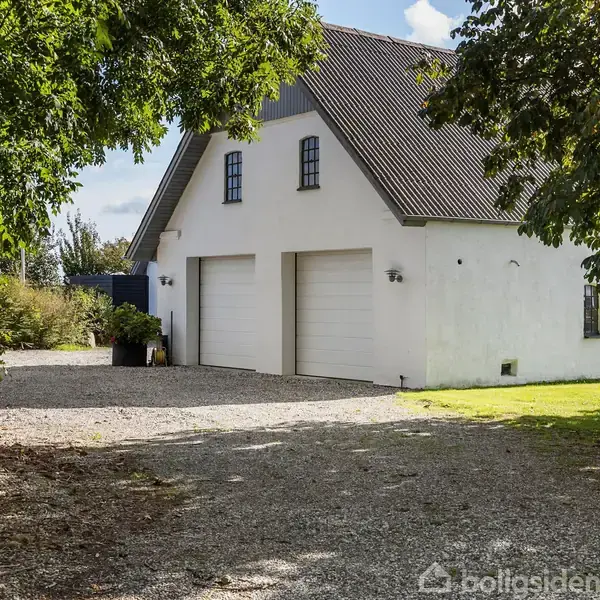 En hvid bygning med to garageporte står stille på en grusindkørsel omgivet af grønne træer og græs. Bygningen har et skråt tag og små vinduer.