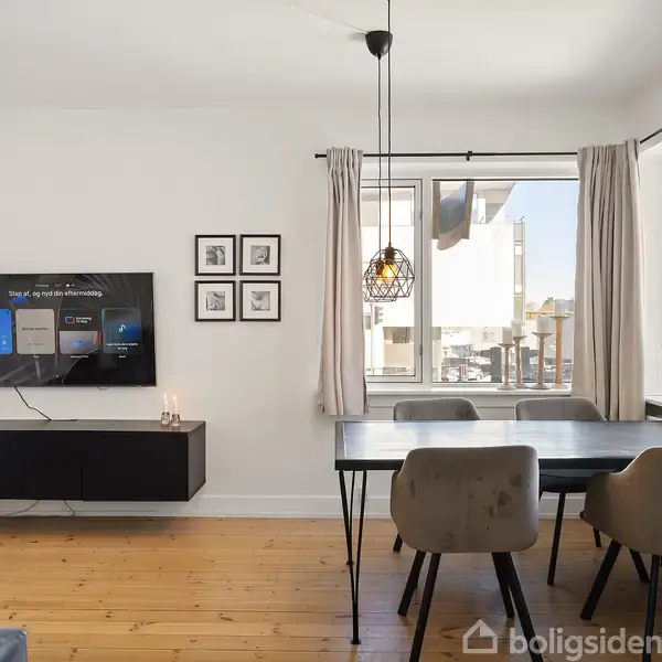 TV hænger på væggen, viser startskærm; stue med moderne indretning, hvide vægge, sorte møbler, store vinduer med lysindfald; spisebord med fire stole, hængende lampe, flere små billeder ved siden af.