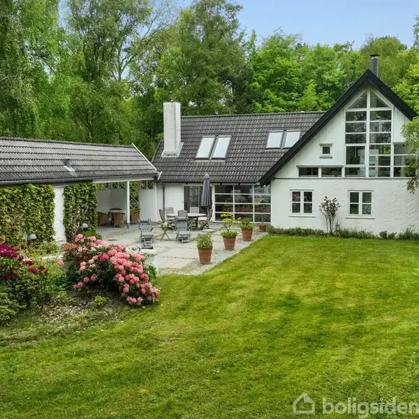 Et hvidt hus med skråtag og store vinduer står i en frodig have med græsplæne og farverige blomster. En terrasse med havemøbler befinder sig mellem huset og en carport.