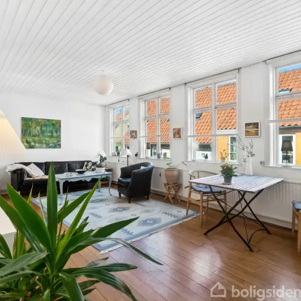 Stue med sofa og lænestol ved siden af et tæppe. Bord og stole står ved store vinduer. Væggene er hvide, med kunst og planter som dekorationer.