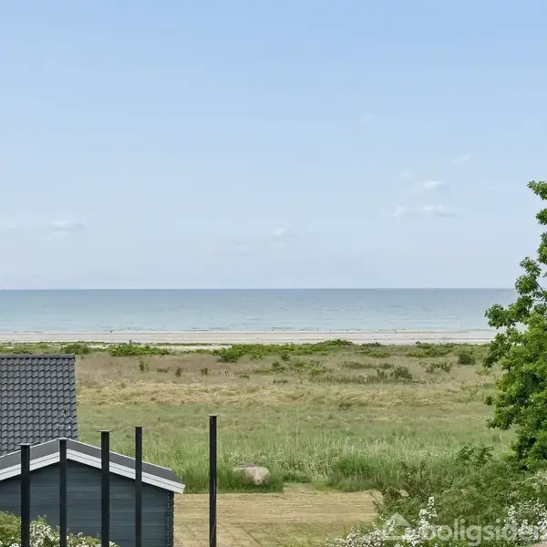 Et hus med gråt tag står foran en græsmark, med hav og sandstrand i baggrunden. En stor grøn busk eller træ er til højre.