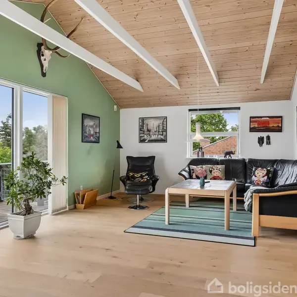 Stue med lædersofa og et sofabord på et tæppe. Rummet har højt, træbeklædt loft og grønne vægge. Store vinduer åbner ud mod en balkon med naturudsigt.