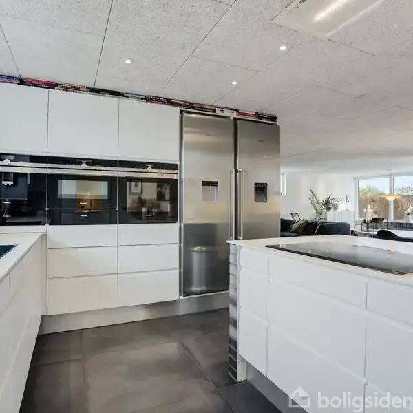 Et moderne køkken med hvide skabe og en integreret ovn ved siden af et stort, rustfrit stål køleskab. Øen har en kogeplade. I baggrunden ses en stue med spisebord.