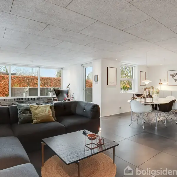 En moderne stue med sort sofa står ved et sofabord med dekorationer. Til højre ses et spisebord med hvide stole og en verdenskort-plakat på væggen. Rummet er lyst med store vinduer.