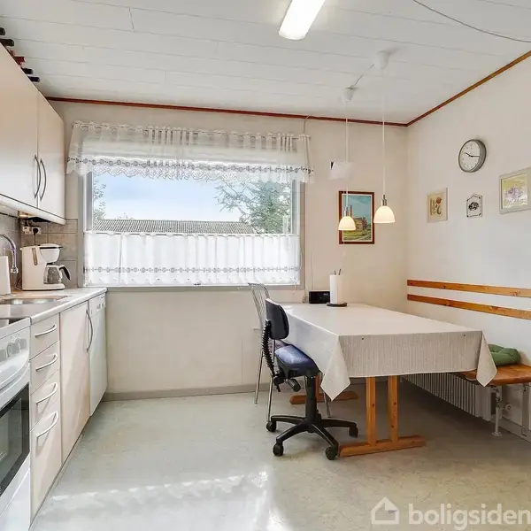 Køkken med lyse skabe, hvor en stol og bord med dug står ved vinduet. Væggene har ure og billeder, og en dør leder til et tilstødende rum.