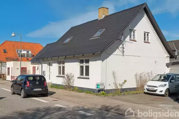 Et hvidt hus med sort tag står langs en vej. To biler er parkeret foran huset. Solrig dag med blå himmel.