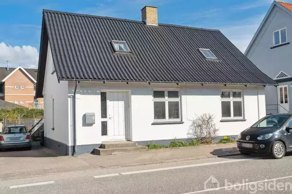 Et hvidt hus med sort tag har en hoveddør og flere vinduer. To biler er parkeret foran huset på en gade i et boligområde.