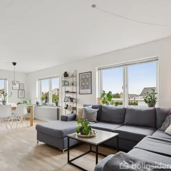 Grå hjørnesofa med puder står i en lys stue, ved siden af et sofabord og foran store vinduer. Spisebord med stole og hængelamper i baggrunden.