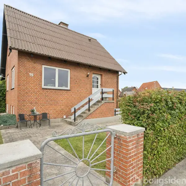 Et murstenshus står med trætrappe og terrasse foran nummer 75. Huset har skråt tag, omgivet af hække og en flisebelagt sti, mens haven er nydeligt indrettet.