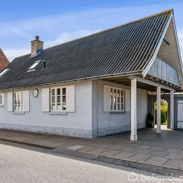 Hus med gråmalet facade og sort tag, står roligt ved en gade. To døre og flere vinduer vender ud mod fortovet. En lille gårdsplads ses ved siden af.
