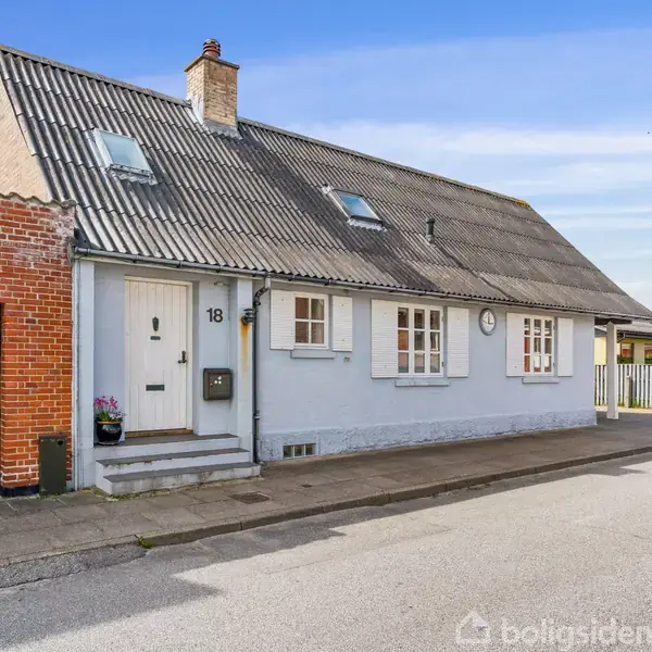 Huset stå stille på en stille gade. Det har en gråt tag og en hvidmalet facade med tre vinduer. Nummeret "18" står ved den hvide dør.