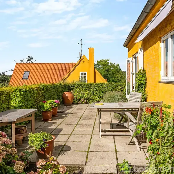 Et bord med stole står på en flisebelagt terrasse foran et gult hus. Omgivet af frodige planter og blomster, ses et andet hus med orange tag i baggrunden.