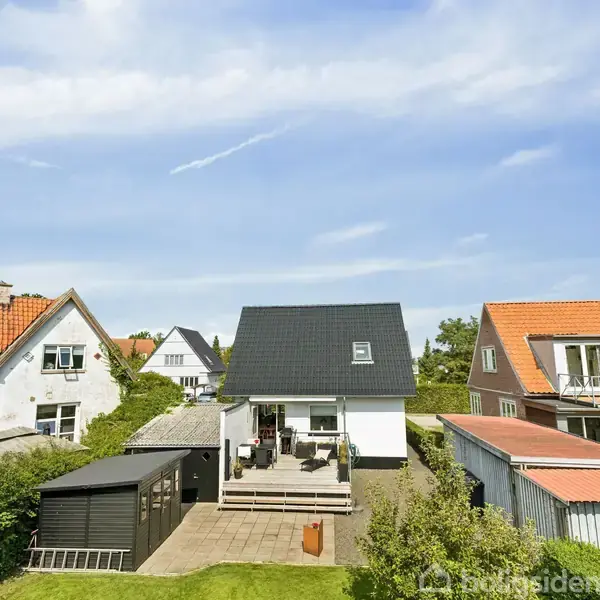 Et hvidt hus med sort tag står i midten, omgivet af to huse med orange tage. En terrasse og have ses foran huset under en blå himmel.