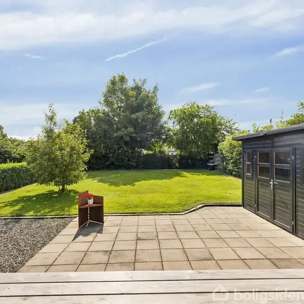 Et sort skur står på en flisebelagt terrasse, mens en rød stol er placeret på grus. Græsplæne og buske omkranser området under en klar blå himmel.
