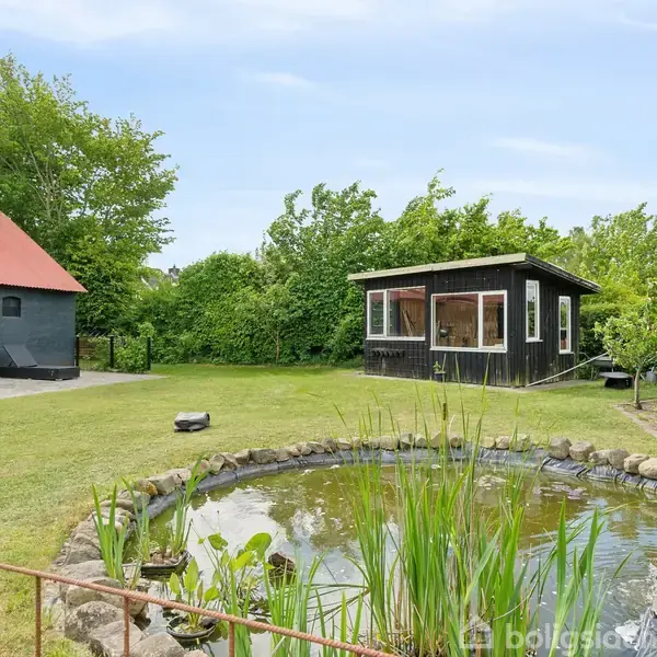 En lille havepavillon står ved siden af en dam omgivet af sten. Bagved ses en bygning med rødt tag, alt sammen i en grøn, frodig have.