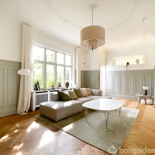 Sofaen står i en lys stue med parketgulv og paneler, omgivet af store vinduer. En moderne lampe hænger over et sofabord, mens en lænestol står i baggrunden.