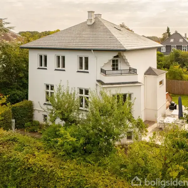 En hvid to-etagers villa står stille omgivet af frodig vegetation med træer og buske, en lille terrasse og en velplejet have i et roligt villakvarter.