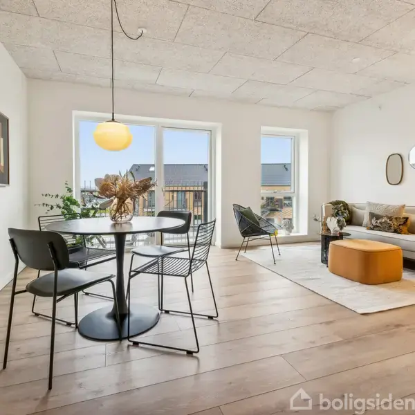 Rundt spisebord med sorte stole står i en lys stue. En sofa med puder og et rundt bord ses ved vinduet. Moderne interiør, træplankegulve.