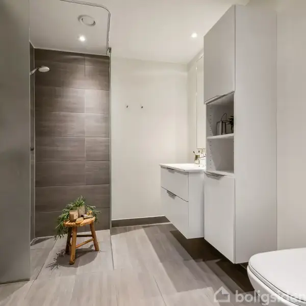 Et moderne badeværelse med en bruser, hvor en stol med en plante står under armaturet. Håndklæder hænger på væggen, og der er et toilet og vaskeskab i rummet.
