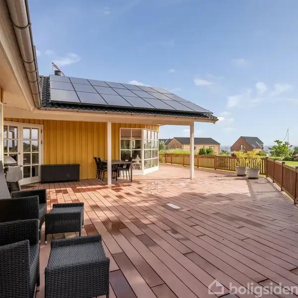 Gult træhus med solpaneler på taget. Stor terrasse med sorte kurvemøbler og bord, omgivet af en lav træhæk. I baggrunden ses grønne græsplæner og nabohuse.