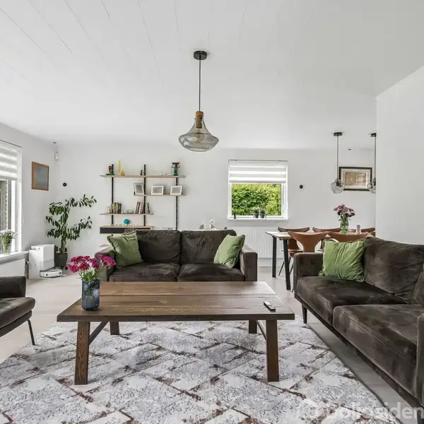 Stuen har to mørke sofaer med grønne puder omkring et træbord. En stor lampe hænger ovenfor. I baggrunden er der et spiseområde og vinduer med udsigt til have.