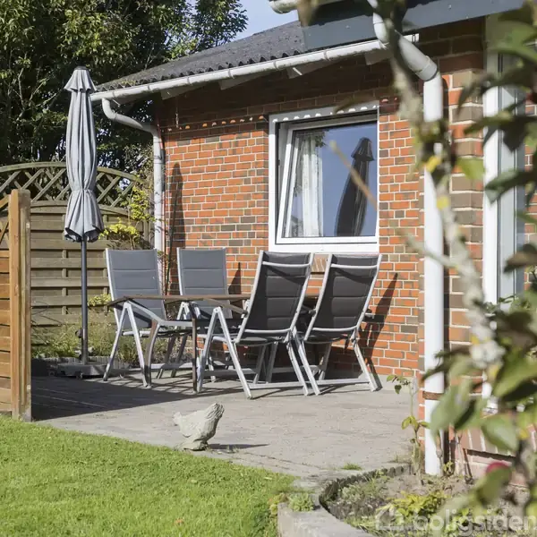 Terrasse med fem sorte stole og parasol står foran et rød murstenshus. Et hegn og græsplæne omkranser området, mens et æbletræ ses i forgrunden og en høne går forbi.