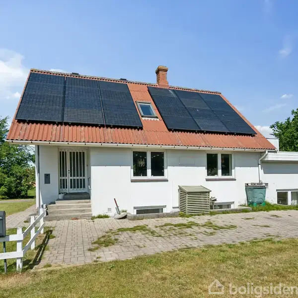 Hus med solpaneler på rødt tag, står på flisebelagt grund omgivet af græsplæne og træer. En postkasse ved hvidt hegn og et til salg-skilt ses ved indkørslen.