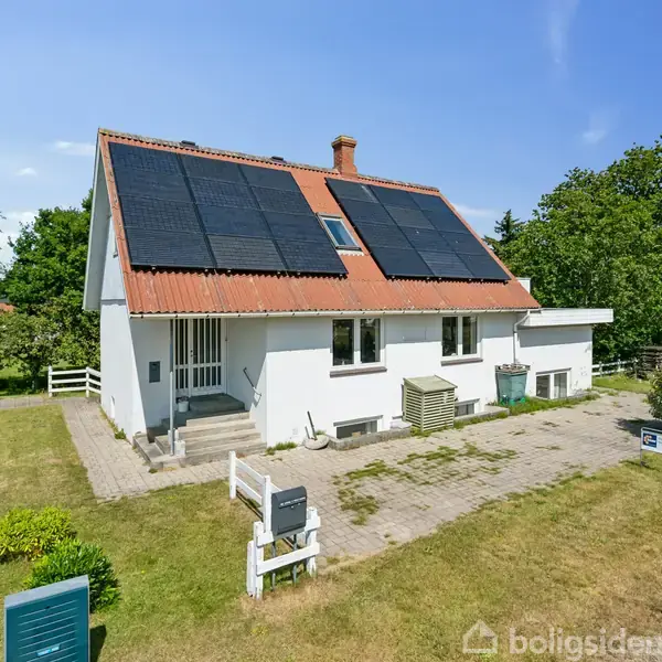 Et hvidt hus med solcellepaneler på taget står stille i en grøn have. Omkring huset er der en græsplæne med buske og en flisebelagt sti.