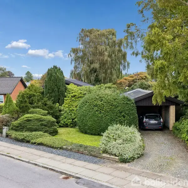 En bil holder parkeret i en carport ved siden af et hus omgivet af frodig have med velplejede buske og træer. Det er en solrig dag med klar blå himmel.