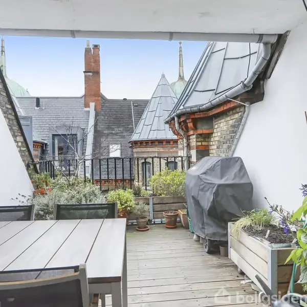 En træterrasse med borde og stole står stille omgivet af planter i krukker. I baggrunden ses tagene fra omkringliggende bygninger, under en åben himmel med let skyer.