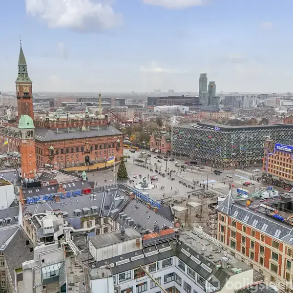 Et bybillede med rådhustårnet som hovedfokus, der rager op i en travl by midt i åbne pladser og omgivende bygninger. Himlen er let overskyet.