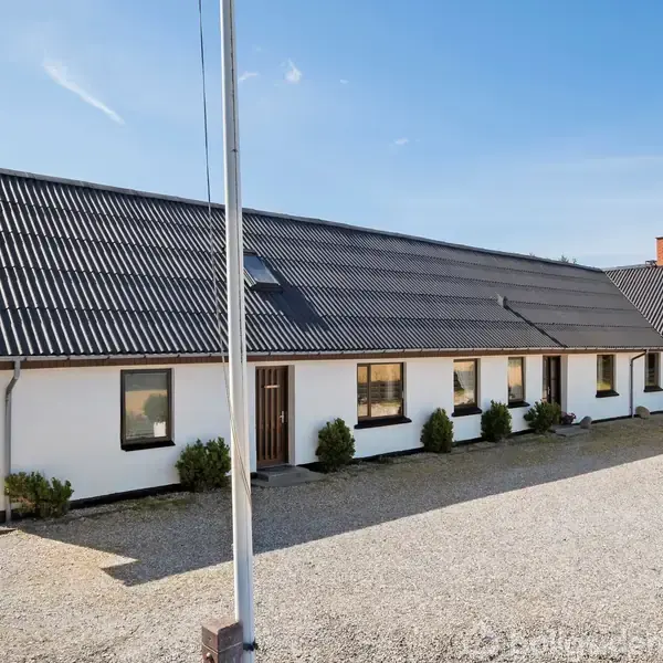 Et hvidt hus med sort tag står stille omgivet af en grusplads og grønne marker. Træer er synlige i baggrunden.