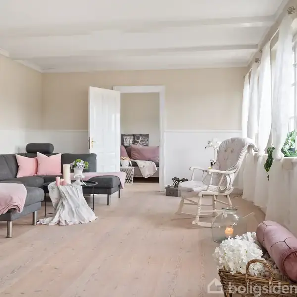 Stue med grå sofa og småborde med lyserøde puder og tæppe. Rocking chair ved vinduer med gardiner. Åben dør viser soveværelse. Kurv med blomster og planter langs vinduet.